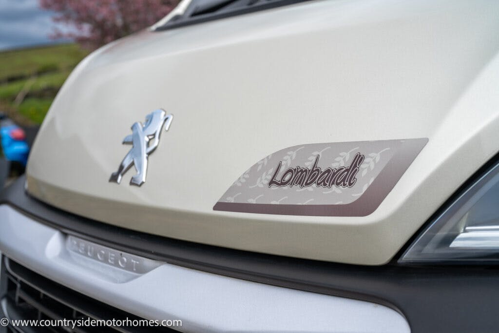 Close-up of a van’s hood featuring the Peugeot emblem and a badge with the word "Lombardi." The URL at the bottom of the image reads "www.countrysidemotorhomes.com." The background shows part of a garden and blurred greenery. This is part of the 2022 Elddis Autoquest Lombardi 150 Masters Collection.