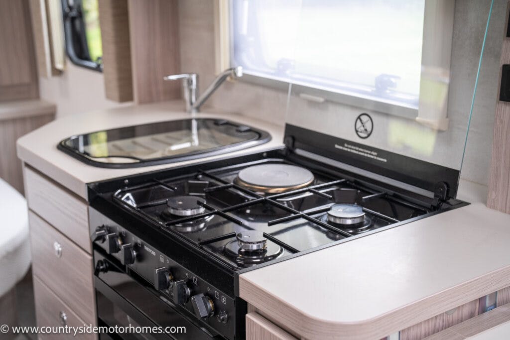 A modern kitchen in the 2022 Elddis Autoquest Lombardi 150 Masters Collection motorhome features a black gas stove and oven with three burners and a small control panel. There's a sink with a faucet adjacent to the stove, plus a window above the countertop that allows natural light to enter.