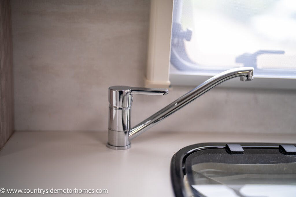 A modern, chrome kitchen faucet with a single handle is installed on a light-colored countertop in the sleek 2022 Elddis Autoquest Lombardi 150 Masters Collection. The faucet is positioned near a window, and a black stove cover is partially visible in the lower right corner.