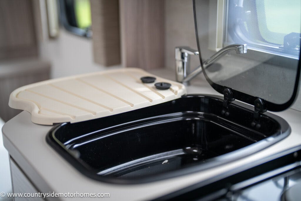 A close-up of a kitchen sink inside the modern 2022 Elddis Autoquest Lombardi 150 Masters Collection motorhome. The black sink has a beige cover, part of which is open, revealing the basin. The faucet is situated behind the sink, and the window above features a partially drawn screen.