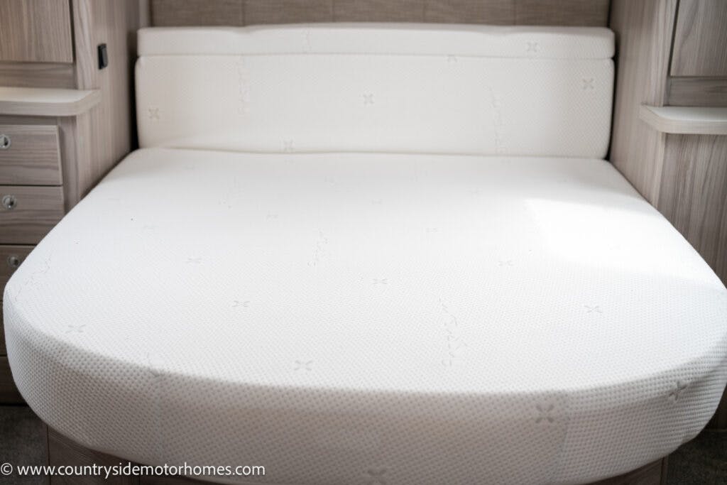 A neatly made round corner bed with a white mattress cover located inside a 2022 Elddis Autoquest Lombardi 150 Masters Collection motorhome. The bed is surrounded by light wooden cabinetry on both sides. Natural light illuminates the bed, casting soft shadows. The website "www.countrysidemotorhomes.com" is visible in the corner.