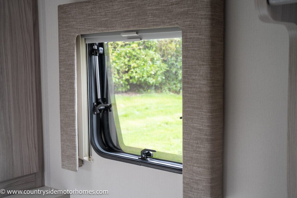 This image showcases a rectangular window in a 2022 Elddis Autoquest Lombardi 150 from the Masters Collection, framed with beige fabric. The window is slightly open, revealing a view of green grass and hedges outside. The interior wall around the window is plain and light-colored.
