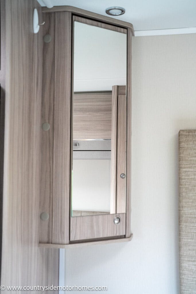 A wooden cabinet with a mirror door is mounted on the wall, reflecting another part of the room. A small portion of the ceiling with a recessed light is visible. The website "www.countrysidemotorhomes.com" promotes the 2022 Elddis Autoquest Lombardi 150 Masters Collection at the bottom.