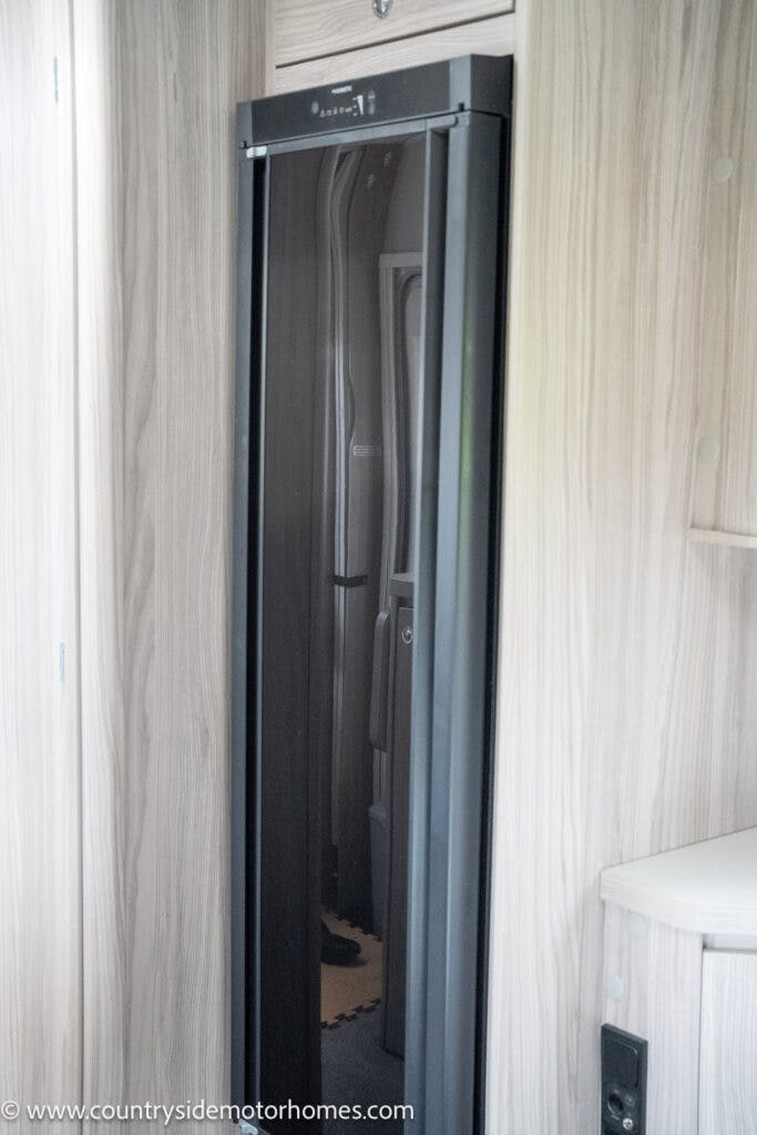 A tall, slim, black refrigerator is installed in a wooden cabinet enclosure inside the 2022 Elddis Autoquest Lombardi 150 Masters Collection RV. The wooden paneling surrounds the appliance, and a portion of a white countertop with a small section of a control panel is visible in the lower right corner.