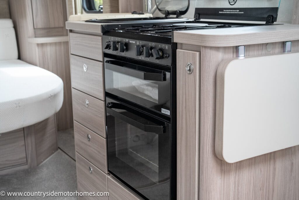A modern kitchen inside the 2022 Elddis Autoquest Lombardi 150 Masters Collection RV showcases a built-in stove with four burners and an oven below, surrounded by light wood cabinetry and drawers. To the right, a fold-down counter extension is visible. The wall panels and flooring are light-colored.