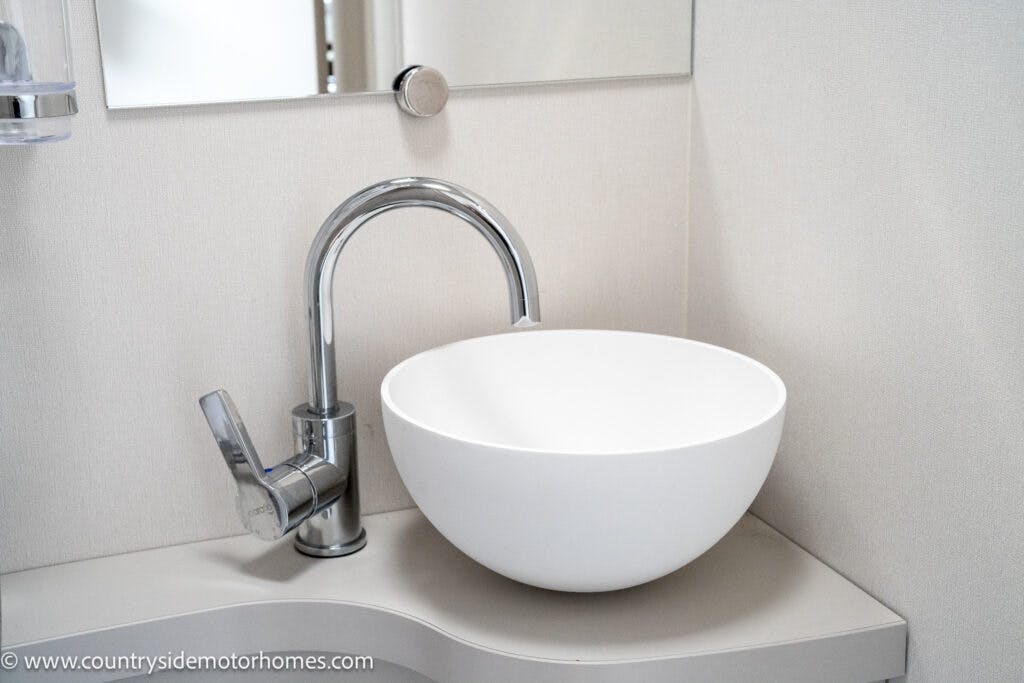 A modern bathroom vanity with a sleek round white sink bowl and a curved chrome faucet, reminiscent of the luxurious finish found in the 2022 Elddis Autoquest Lombardi 150 Masters Collection. The sink is positioned on a light gray countertop, and a mirror is mounted on the wall behind it, maintaining a clean and minimalist design.