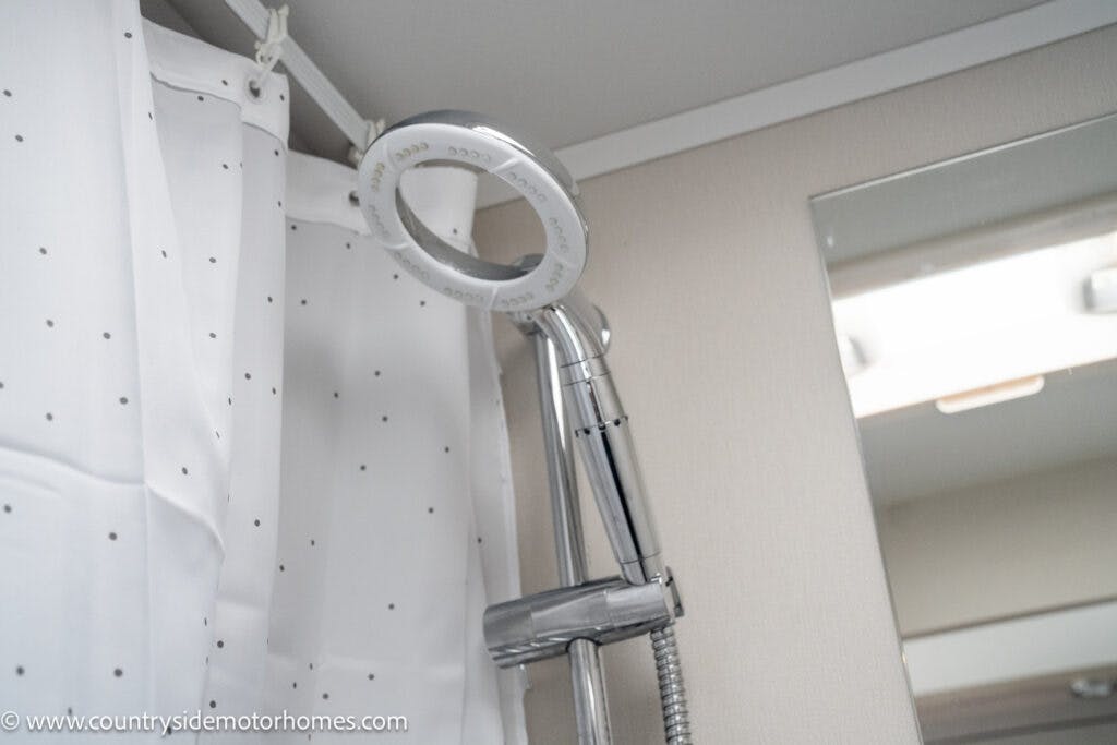 A close-up view of a showerhead with a circular design attached to a metal hose in the 2022 Elddis Autoquest Lombardi 150 Masters Collection. The showerhead is installed in a shower area with a white curtain and a small portion of a mirror visible in the background.