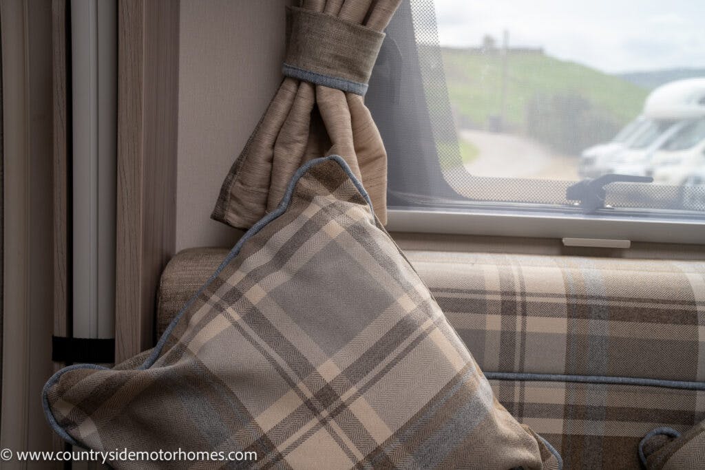 An interior view of the 2022 Elddis Autoquest Lombardi 150 Masters Collection motorhome features a plaid cushion and matching curtain tie near a window. Through the window, part of another motorhome and a slightly blurred view of the outdoors are visible.