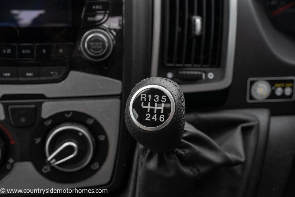 Close-up image of a gear stick in a 2022 Elddis Autoquest Lombardi 150 Master's Collection. The gear pattern shows six forward gears and reverse. Surrounding details include part of the dashboard controls, buttons, and a section of the air conditioning system. The website "www.countryside motorhomes.com" is visible at the bottom.