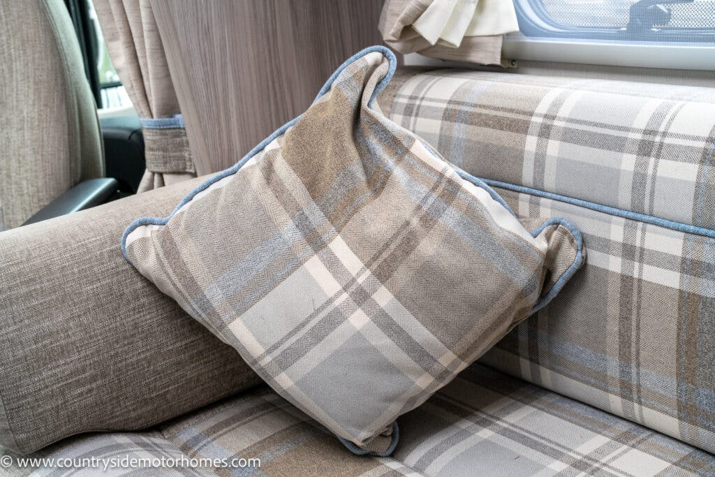 A grey and beige plaid cushion with blue piping is placed on a beige upholstered seat inside a 2022 Elddis Autoquest motorhome. In the background, there are curtains, a part of a window, and a section of the motorhome's interior wall from the Lombardi 150 Masters Collection.