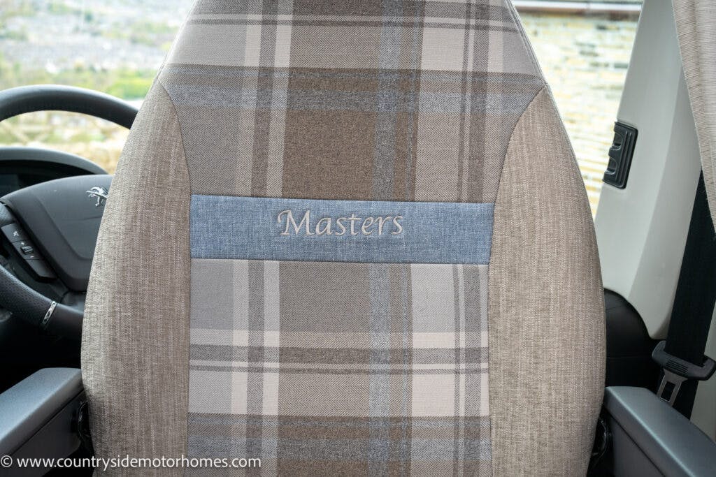 Close-up of a car seat with a plaid fabric pattern in the 2022 Elddis Autoquest Lombardi 150 Masters Collection. The word "Masters" is embroidered on a horizontal blue strip in the center of the seat's backrest. The steering wheel and part of the dashboard are visible on the left side of the image.