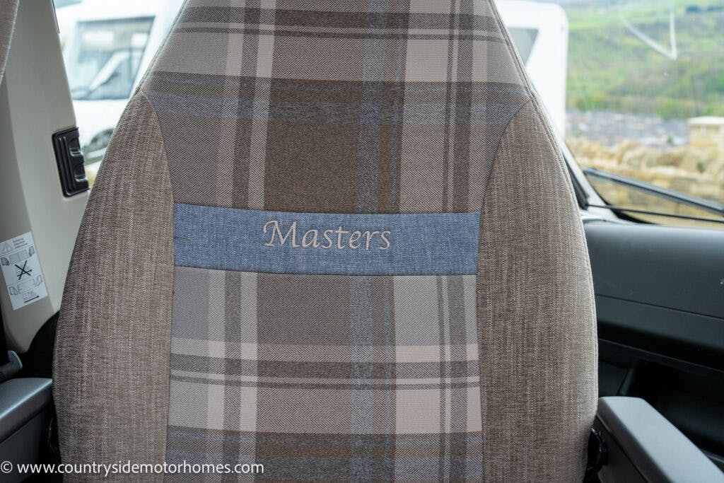 A close-up of a fabric seat with a plaid pattern in brown, gray, and beige tones from the 2022 Elddis Autoquest Lombardi 150 Masters Collection. The headrest features a blue strip with "Masters" embroidered in white. The seat is in a vehicle with a window in the background. CountrysideMotorhomes.com is visible in the lower-left corner.