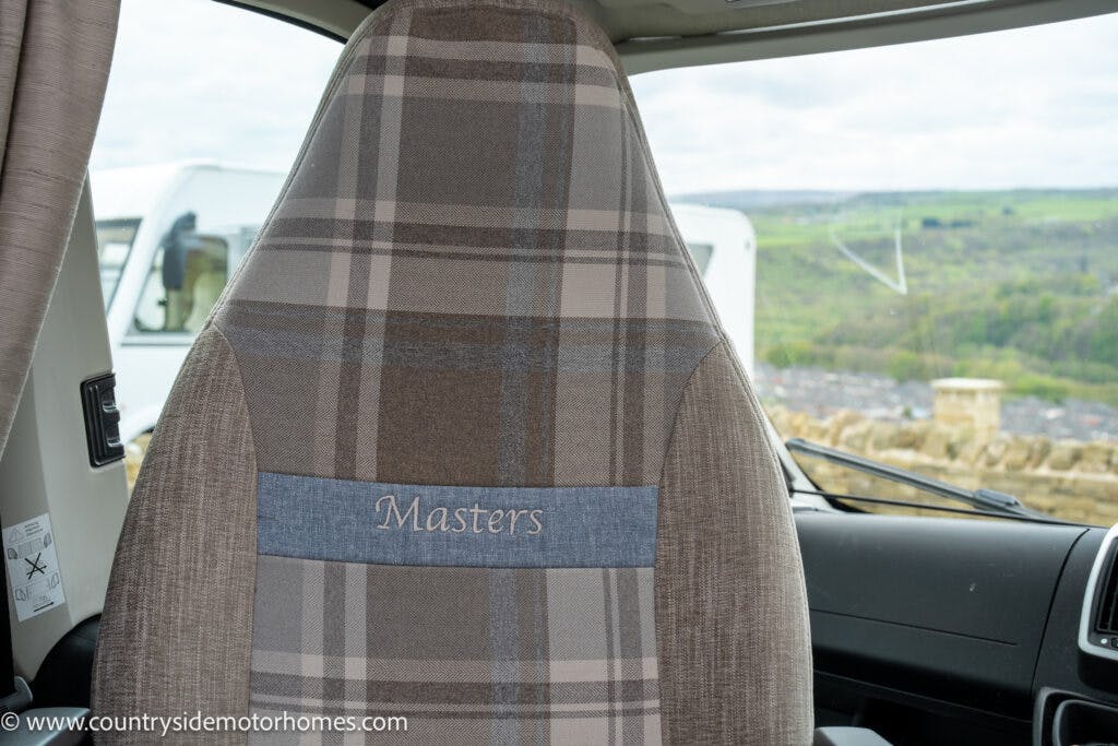 The image shows the backrest of a driver's seat in a 2022 Elddis Autoquest Lombardi 150 from the Masters Collection. The seat has a plaid fabric design with the word "Masters" embroidered on a blue panel. The windshield reveals a parked RV and a scenic, hilly landscape in the background.