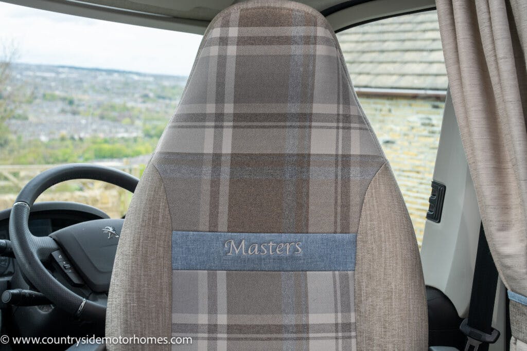 Photograph showing the driver's seat of a 2022 Elddis Autoquest Lombardi 150 Masters Collection motorhome, upholstered in a beige and gray plaid fabric with the word "Masters" embroidered on a blue panel in the center. The steering wheel and part of the dashboard are visible to the left.