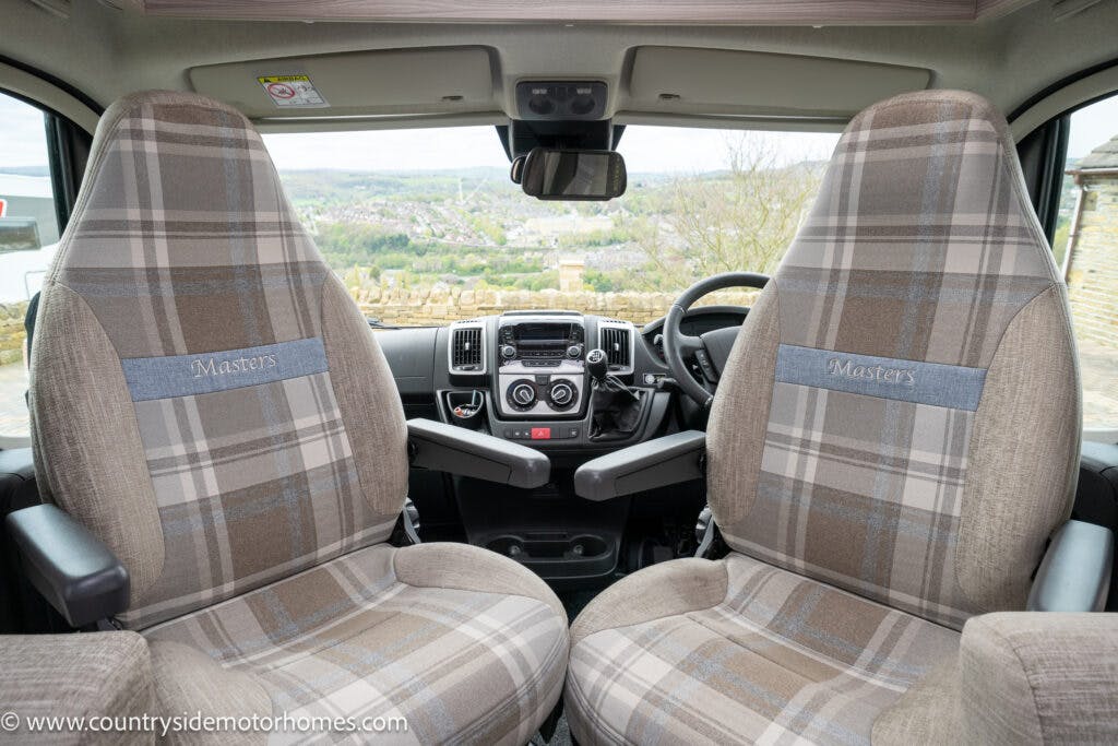The interior front seating area of the 2022 Elddis Autoquest Lombardi 150 Masters Collection motorhome shows two plaid-patterned captain's chairs with “Masters” embroidered on the headrests. The dashboard with steering wheel, gearshift, and controls is visible through the gap between the seats.