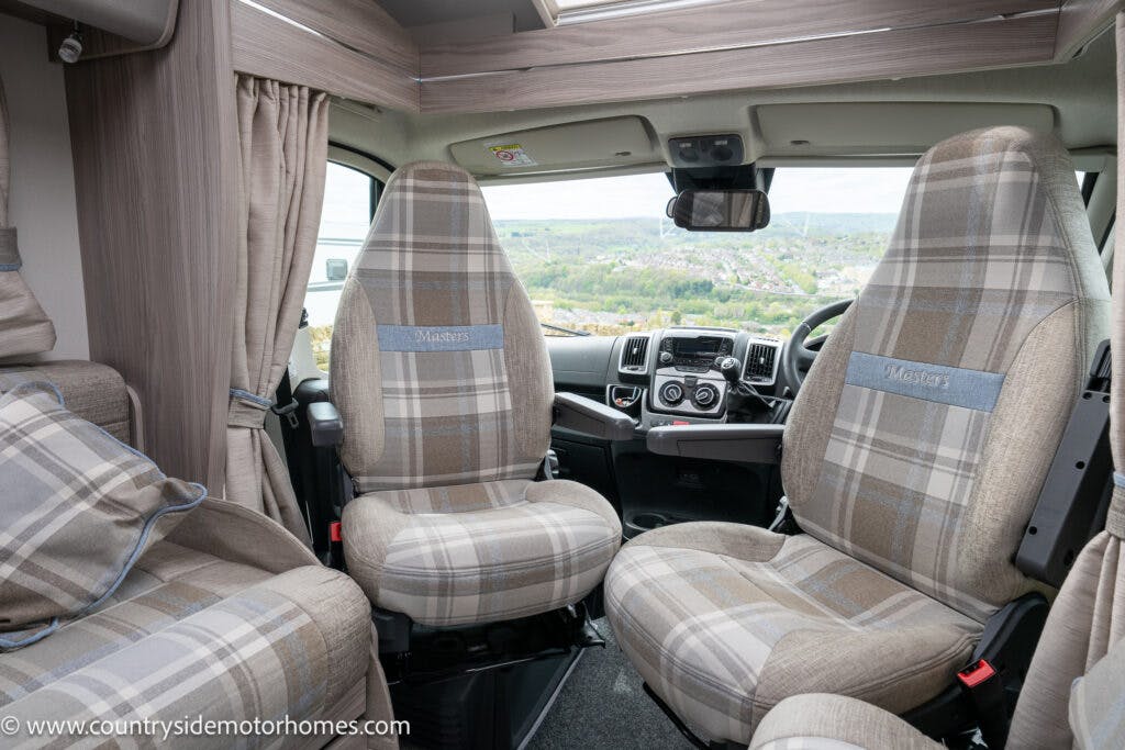 The image shows the interior of a 2022 Elddis Autoquest Lombardi 150 motorhome, with two front seats upholstered in plaid fabric and branded with "Master's." The dashboard and steering wheel are visible, and there is a view of a landscape through the front windshield. Curtains are partially closed.