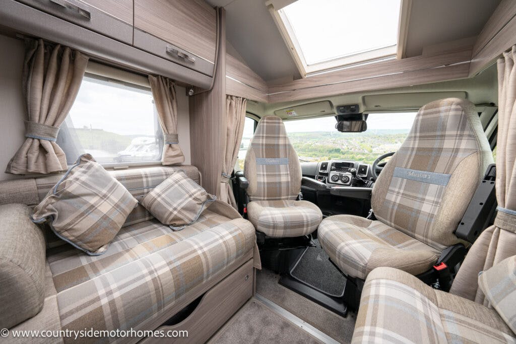Interior of the 2022 Elddis Autoquest Lombardi 150 Masters Collection motorhome showing the front cabin with two plaid-patterned seats, a dashboard, and a large windshield. The living area includes a matching plaid sofa and curtained windows on both sides, with overhead storage compartments and a skylight.