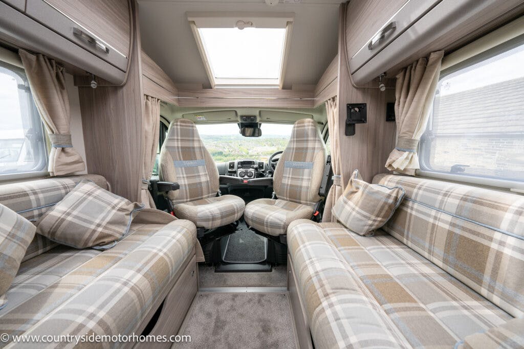The interior of the 2022 Elddis Autoquest Lombardi 150 Masters Collection motorhome features two front seats, a skylight, and a lounge area with plaid upholstery on the sofas. The space is bright with beige-toned walls, windows covered with matching plaid curtains, and a view of the front dashboard.