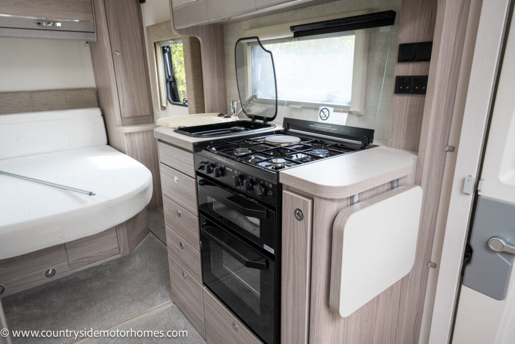 The image shows the interior of a 2022 Elddis Autoquest Lombardi 150 motorhome kitchen. There is a stove with four burners and an oven. Above the stove is a clear, hinged cover. Cabinets and drawers line the area. To the left, part of a bed is visible. The floor is carpeted in this Masters Collection model.