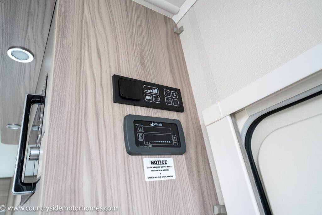 A wall-mounted control panel with various buttons and a small screen, alongside another digital display, in an enclosed space with light wood paneling and a notice card below. A partial view of an overhead cupboard on the left complements the features of the 2022 Elddis Autoquest Lombardi 150 Masters Collection.