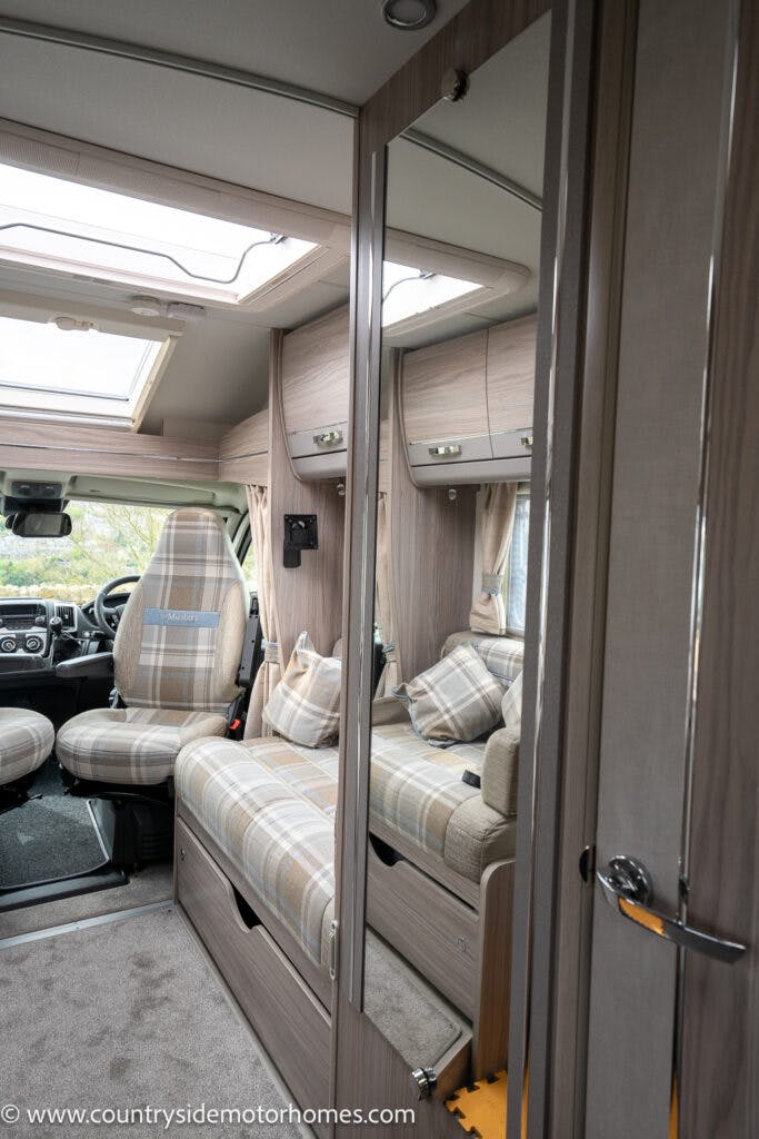 Interior of the 2022 Elddis Autoquest Lombardi 150 Masters Collection, featuring plaid seats and a convertible sofa. The area is equipped with a steering wheel, a dashboard, overhead storage compartments, and a full-length mirror on a wooden wardrobe door. The space has a modern, compact design.