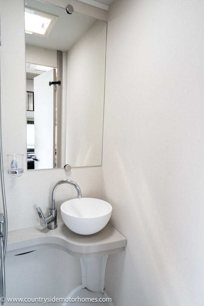 A compact bathroom in the 2022 Elddis Autoquest Lombardi 150 Masters Collection features a round white sink with a silver faucet set on a small L-shaped countertop, underneath a rectangular mirror. The light beige walls complement the sleek design, and a small soap dish is mounted next to the sink.
