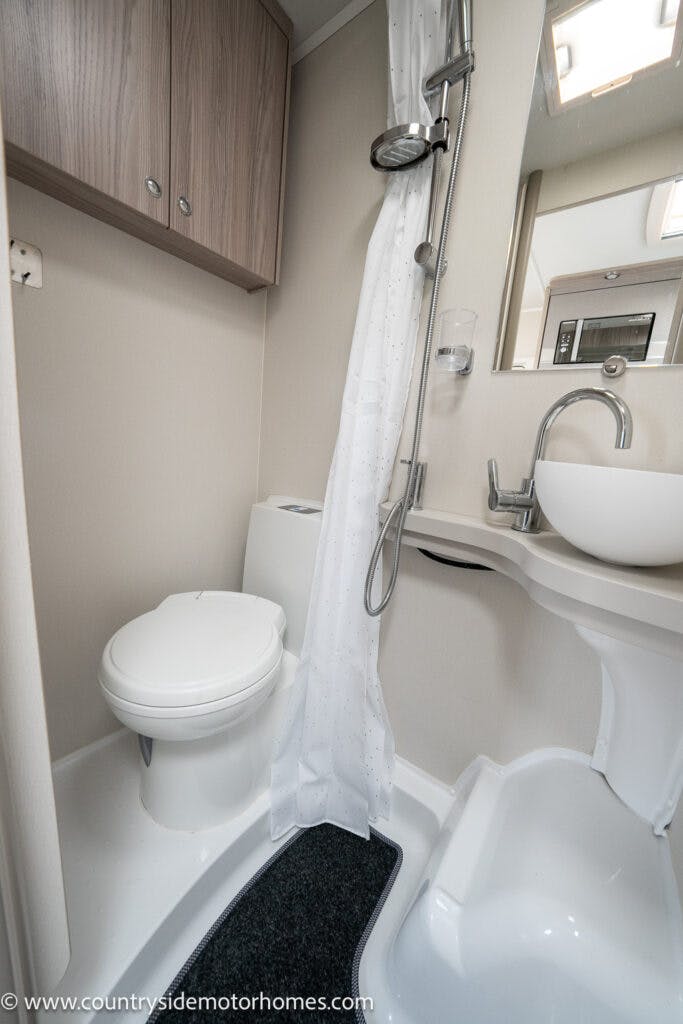 A compact bathroom inside the 2022 Elddis Autoquest Lombardi 150 Masters Collection motorhome features a toilet, a small sink, a mirror, and a shower area with a white shower curtain. The walls and cabinetry are light-colored, and there's a black mat on the floor.