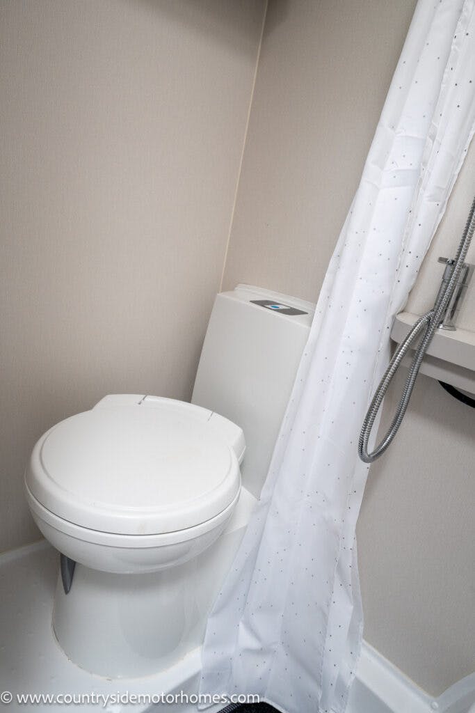 A small bathroom in the 2022 Elddis Autoquest Lombardi 150 Masters Collection with a white toilet, a shower featuring a white curtain, and a hose attached to the wall. The light-colored floor and walls enhance the clean environment. Image from countrysidemotorhomes.com.