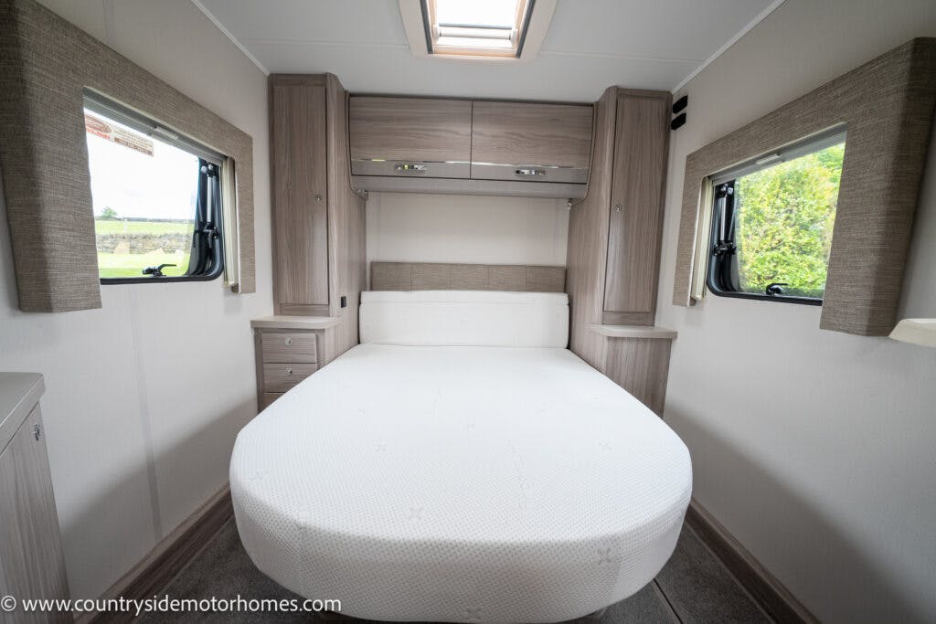 Interior of the 2022 Elddis Autoquest Lombardi 150 Masters Collection motorhome bedroom with a double bed centered against the back wall. The room has two side windows with curtains, overhead cabinets above the bed, and a small skylight. The walls and cabinetry are in neutral tones. A URL is visible in the bottom left corner.