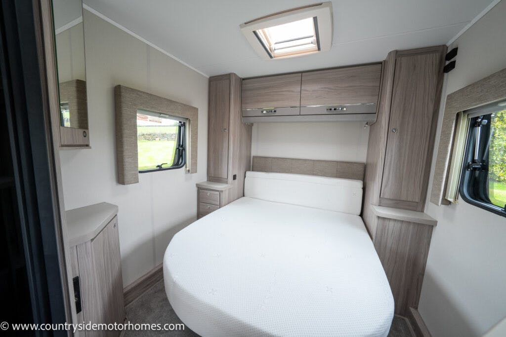 Interior of the 2022 Elddis Autoquest Lombardi 150 Masters Collection motorhome bedroom featuring a double bed with white bedding, overhead storage cabinets, wall-mounted mirrored cabinets on either side, and a built-in bedside cabinet. There is a small window on the right side and a skylight above.