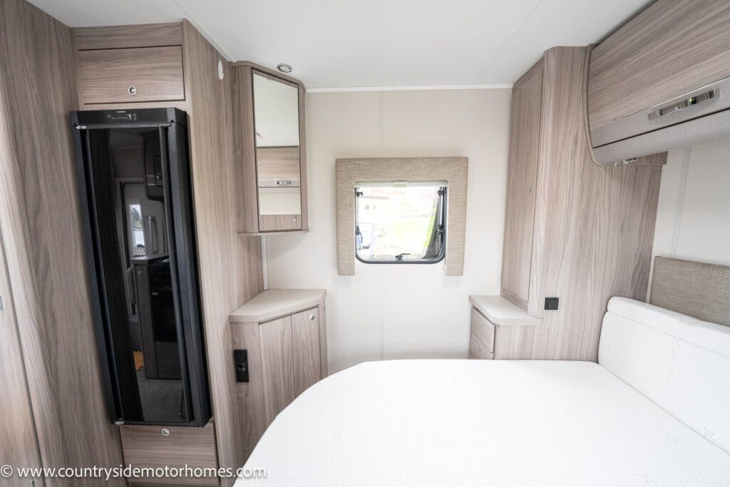 Interior view of the 2022 Elddis Autoquest Lombardi 150 Masters Collection motorhome bedroom featuring a white mattress, wooden cabinets, a small rectangular window, an air conditioning unit above the bed, and a small nightstand. The space is compact and organized with light-colored wood finishes.
