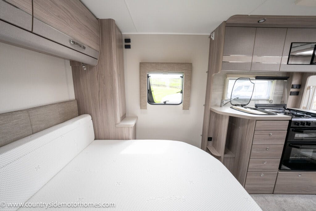 The image shows the interior of a 2022 Elddis Autoquest Lombardi 150 Masters Collection motorhome. It features a compact living space with a bed on the left, a central window with a wooden frame, and a kitchenette on the right with stove, oven, sink, and cabinets. The interior has a modern design.
