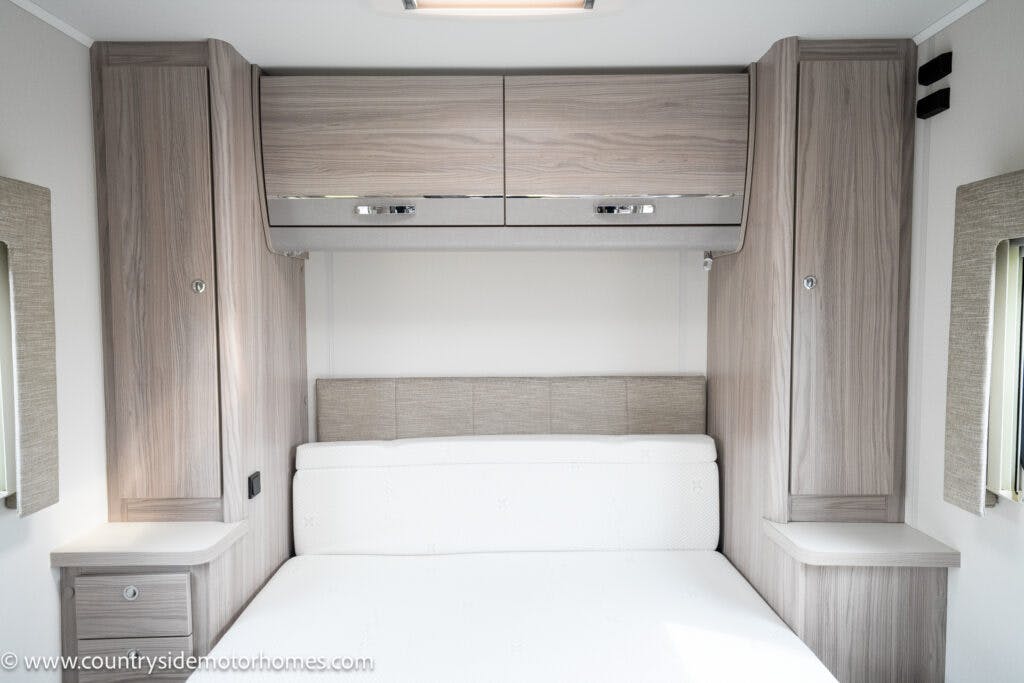 The image shows the interior of a 2022 Elddis Autoquest Lombardi 150 Masters Collection RV bedroom. It features a double bed with a wooden headboard and two bedside tables. Above the bed, there are two wooden storage cabinets and small rectangular windows on both sides.