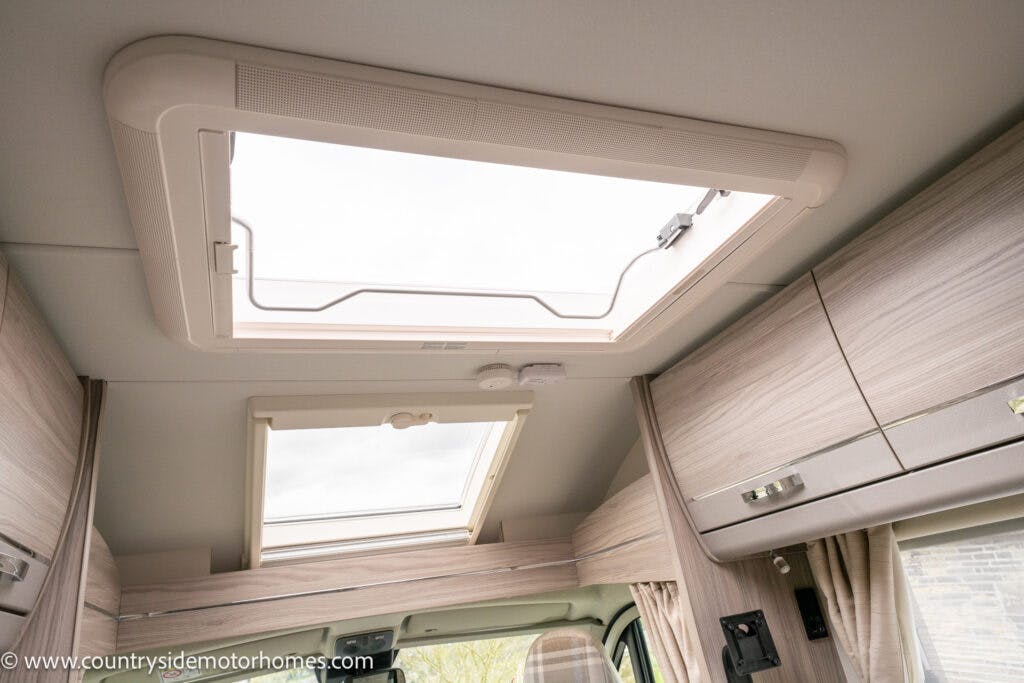Interior view of the 2022 Elddis Autoquest Lombardi 150 Masters Collection motorhome showing an open sunroof on the ceiling, wooden cabinets, a window with a curtain beside the driver's seat, and a beige interior finish. The website URL "www.countrysidemotorhomes.com" is visible on the bottom-left corner.