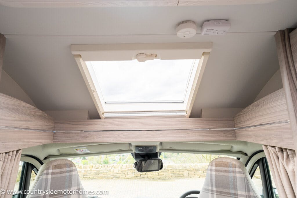 Interior view of the 2022 Elddis Autoquest Lombardi 150 Masters Collection motorhome featuring a skylight on the ceiling, allowing natural light into the vehicle. Seats with checkered upholstery are visible at the front. Wooden cabinetry surrounds the skylight. A smoke detector and control panel are mounted on the ceiling.