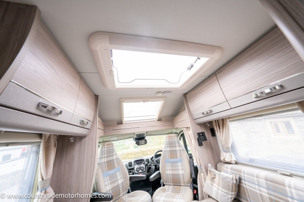 The image shows the interior of a 2022 Elddis Autoquest Lombardi 150 Masters Collection motorhome. It features two front seats with plaid fabric, large windows with blinds, and overhead storage cabinets. A sunroof is present on the ceiling, and part of the driver's dashboard is visible.