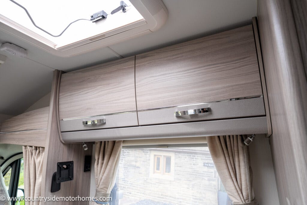 A close-up view of overhead cabinets in the 2022 Elddis Autoquest Lombardi 150 Masters Collection motorhome. The cabinets are wooden with sleek, chrome handles. Below them is a window with beige curtains, providing a view of a stone building outside. An open skylight is visible on the ceiling.