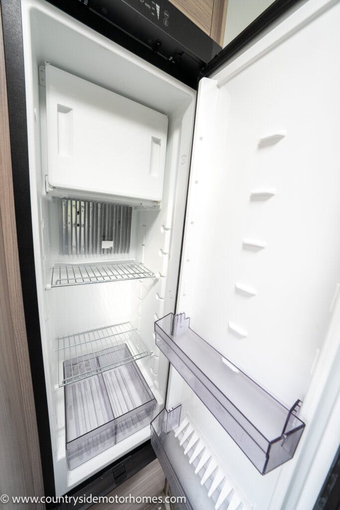 An open, empty refrigerator with a freezer compartment at the top, reminiscent of the streamlined efficiency found in the 2022 Elddis Autoquest Lombardi 150 Masters Collection. The fridge boasts three wire shelves and two transparent plastic shelves on the door. The interior is clean and polished in a sleek white finish.