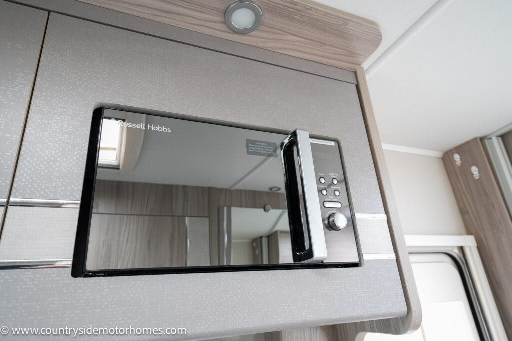 A 2022 Elddis Autoquest Lombardi 150 Masters Collection boasts a microwave oven with a stainless steel front mounted in a wooden cabinet. The microwave door has a reflective finish, showing part of the surrounding interior. The brand name "Russell Hobbs" is visible on the top left corner of the microwave.