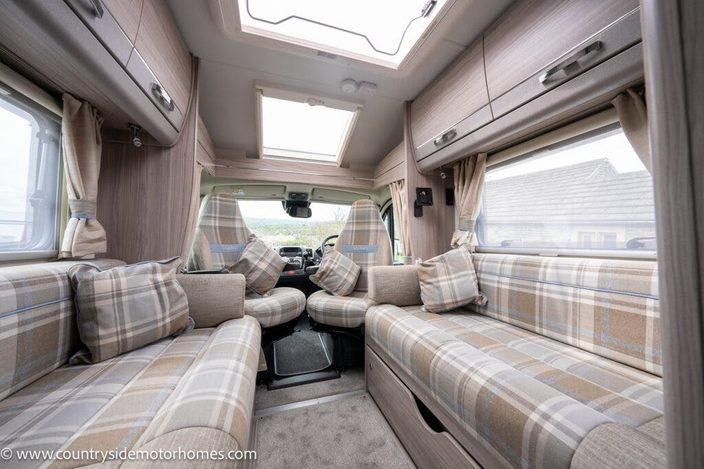 The interior of the 2022 Elddis Autoquest Lombardi 150 Masters Collection motorhome features plaid-patterned seats and cushions, wide windows with beige curtains, and overhead storage cabinets. A sunroof is visible above, providing natural light. The driver's area is in the distance, and the floor is carpeted.