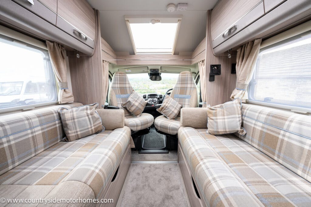 The image shows the interior of a 2022 Elddis Autoquest, featuring a seating area with plaid cushions and beige upholstery. The front cab has two swivel seats facing towards the back. The space is well-lit by windows and a skylight, while storage cabinets are above the seats in this Lombardi 150 Masters Collection motorhome.