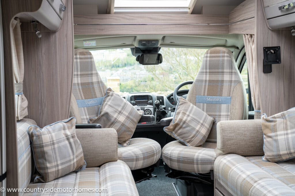 Interior view of the 2022 Elddis Autoquest Lombardi 150 Masters Collection camper van featuring plaid fabric seats and cushions. The front has two captain chairs facing forward with large windows. The dashboard includes a steering wheel, control panel, and display screen. Walls and ceiling are covered in light wood panels.