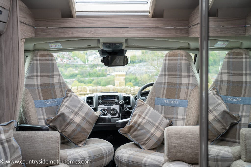 The image captures the interior of a 2022 Elddis Autoquest Lombardi 150 Masters Collection motorhome from the rear-facing front seats. The seats and adjacent cushions are upholstered in a plaid pattern, and you can see the dashboard and steering wheel with a scenic view outside the windshield.