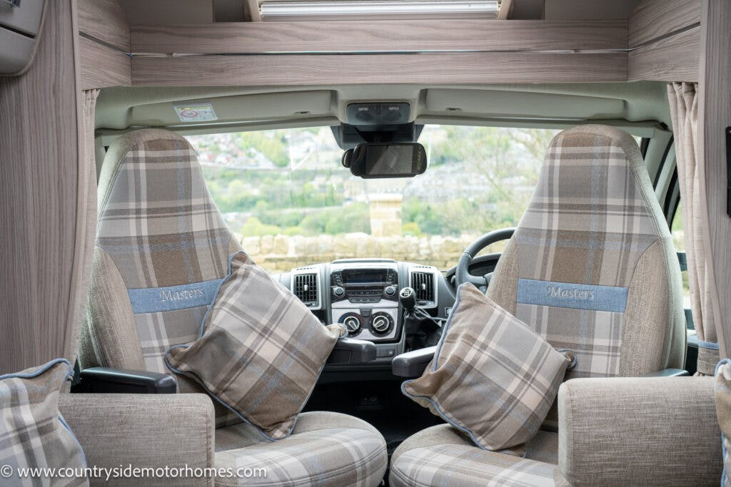 The image shows the interior front view of a 2022 Elddis Autoquest Lombardi 150 Masters Collection motorhome, featuring two plaid-patterned seats with matching pillows. The dashboard includes a steering wheel, control panel, and two cup holders. A large windshield offers a view of the outdoors.