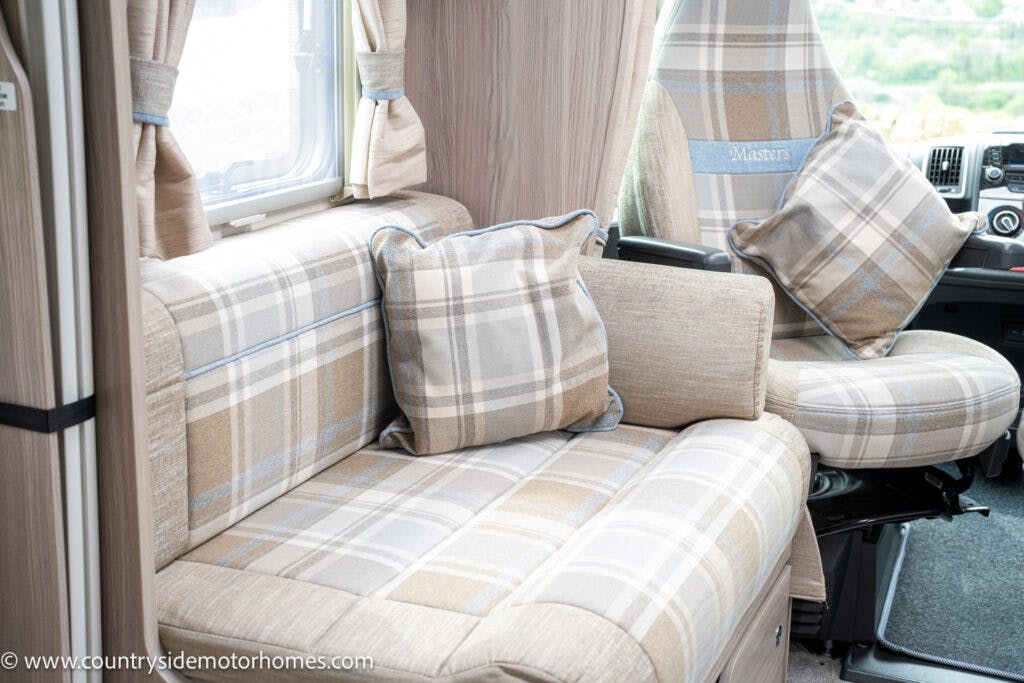 Interior of the 2022 Elddis Autoquest Lombardi 150 featuring a plaid upholstered couch with matching cushions and a driver's seat in similar plaid fabric. A small window with beige curtains is above the couch. The dashboard and part of the steering wheel are visible, showcasing the Masters Collection design.