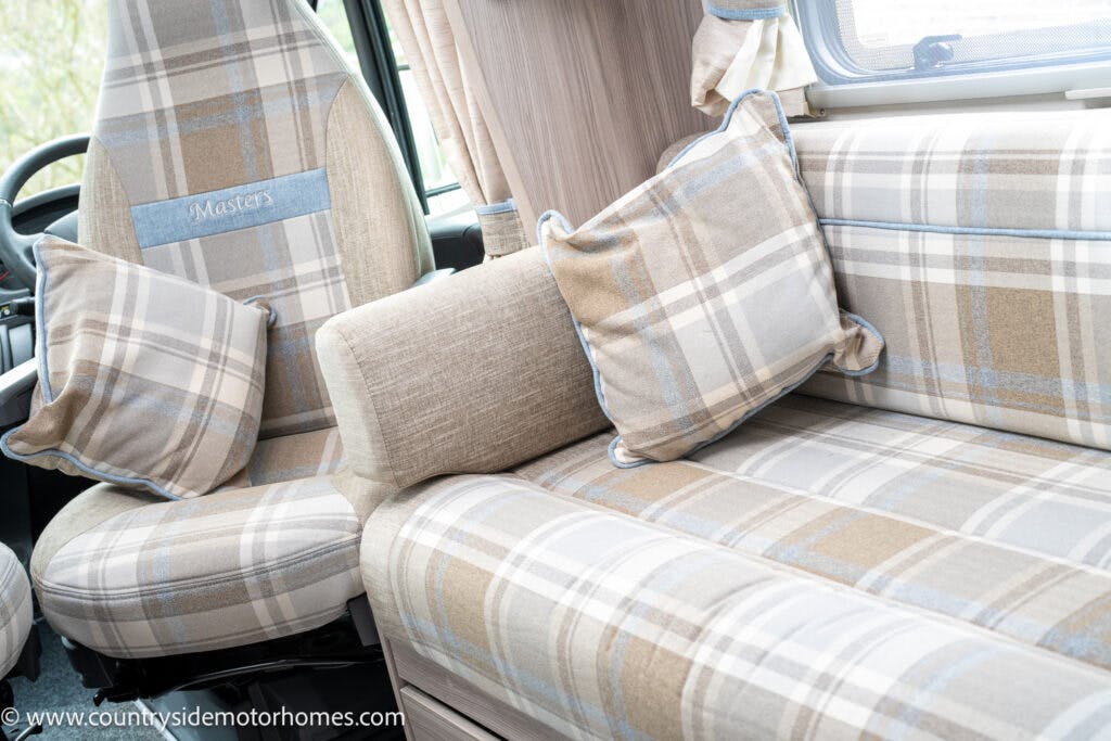 The image shows the interior of a 2022 Elddis Autoquest Lombardi 150 Masters Collection camper van, focusing on the plaid-patterned upholstery of the seating area and driver's seat. There are two matching pillows and a bolster on the banquette, with upholstery in beige, gray, and white tones.