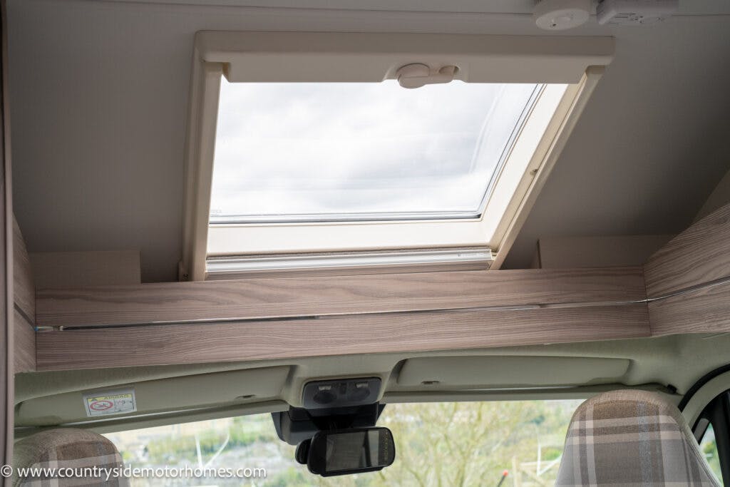 The image shows the interior view of a 2022 Elddis Autoquest Lombardi 150 Masters Collection motorhome, focusing on a roof skylight above the driver's area. The skylight is set in a wooden frame with a light finish, and there is a plaid-patterned headrest visible in the lower right corner. The URL www.countrysidemotorhomes.com is present in the