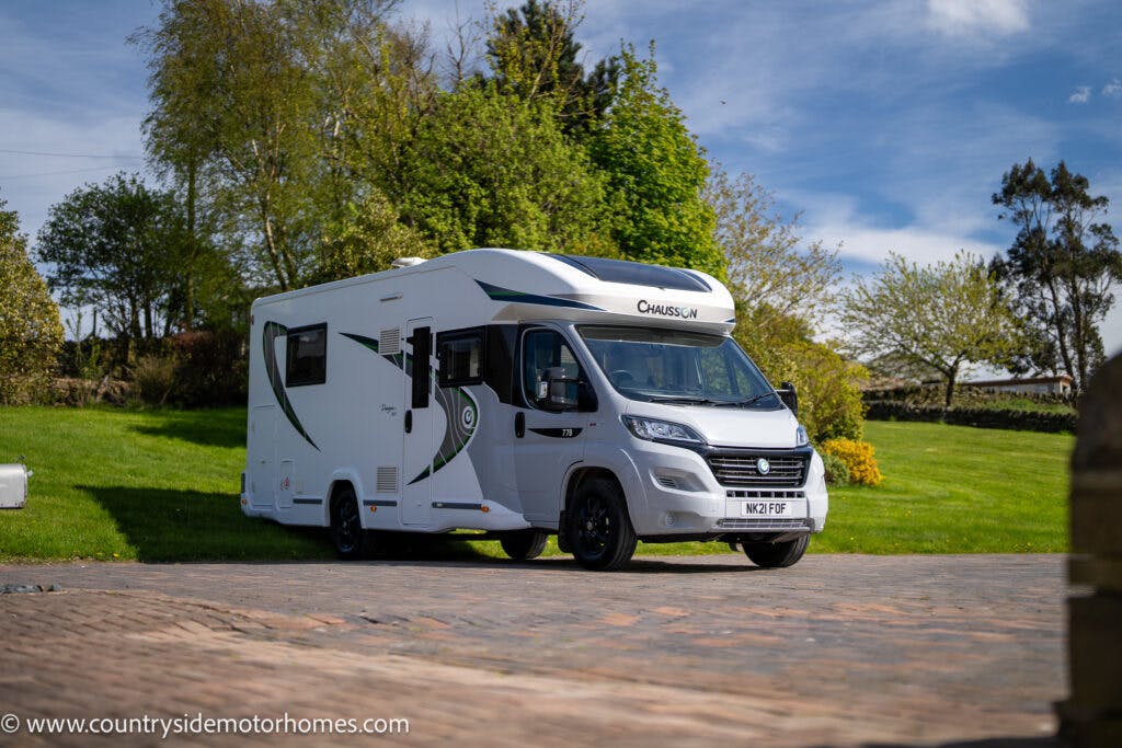 A white 2021 Chausson 778 Premium motorhome with green accents is parked on a paved area with trees and grass in the background. Branded "Chausson," the motorhome's license plate reads "NG21 PGF." A website URL is visible at the bottom of the image.