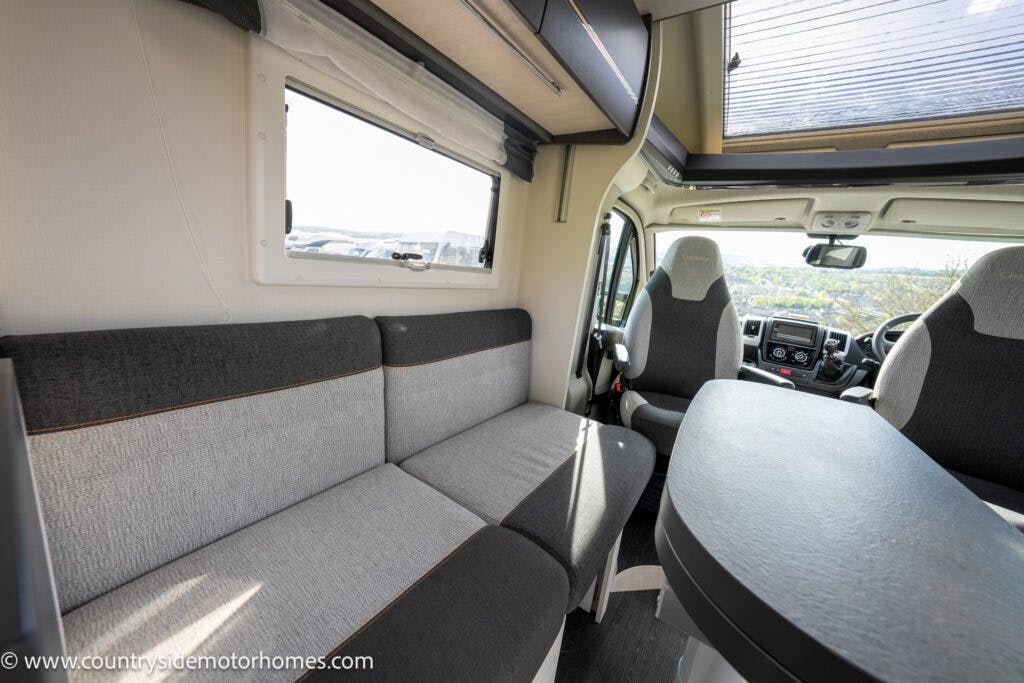 Interior of the 2021 Chausson 778 Premium motorhome featuring a grey and black upholstered sofa, a round table, and the driver's cabin with two seats. The window on the left provides a view of the outside, and sunlight is coming through the windshield.