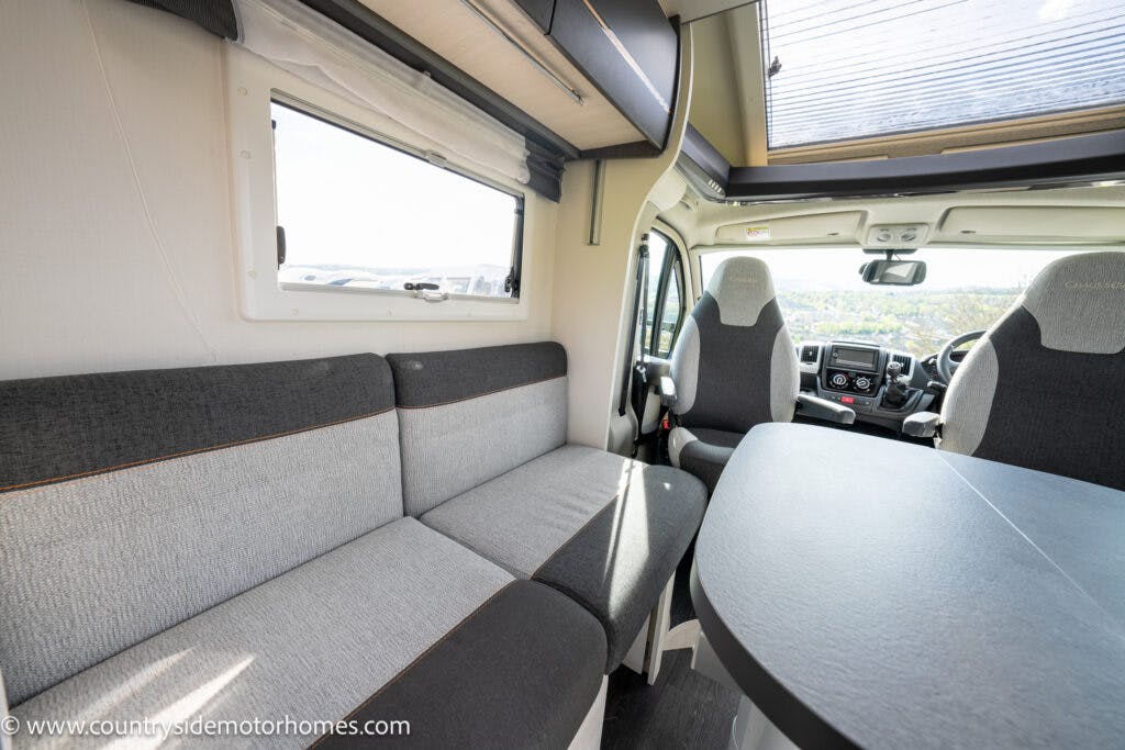 The interior of the 2021 Chausson 778 Premium motorhome features a spacious seating area with gray upholstered cushions, a round table, two front seats, and large windows with blinds. The image is from Countryside Motorhomes, as noted in the URL watermark.
